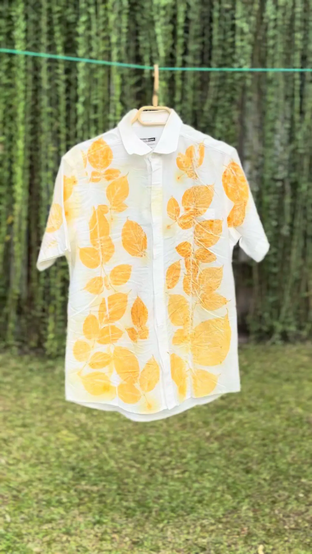 A white shirt printed with golden leaf impressions sways gently on a washing line in front of a wall of green vines.