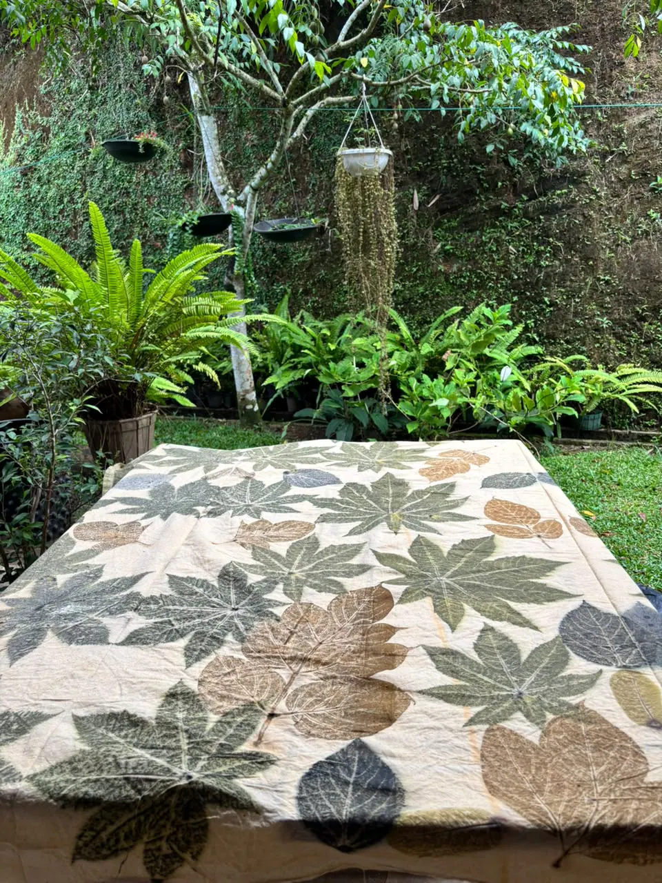 The palmate-leaf cloth spread over a table in a lush garden of ferns and potted plants.