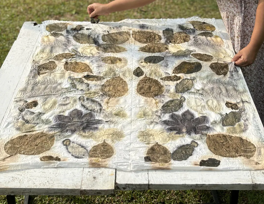 Hands lifting steamed leaves from a cloth printed in umber, olive and charcoal on an outdoor table.